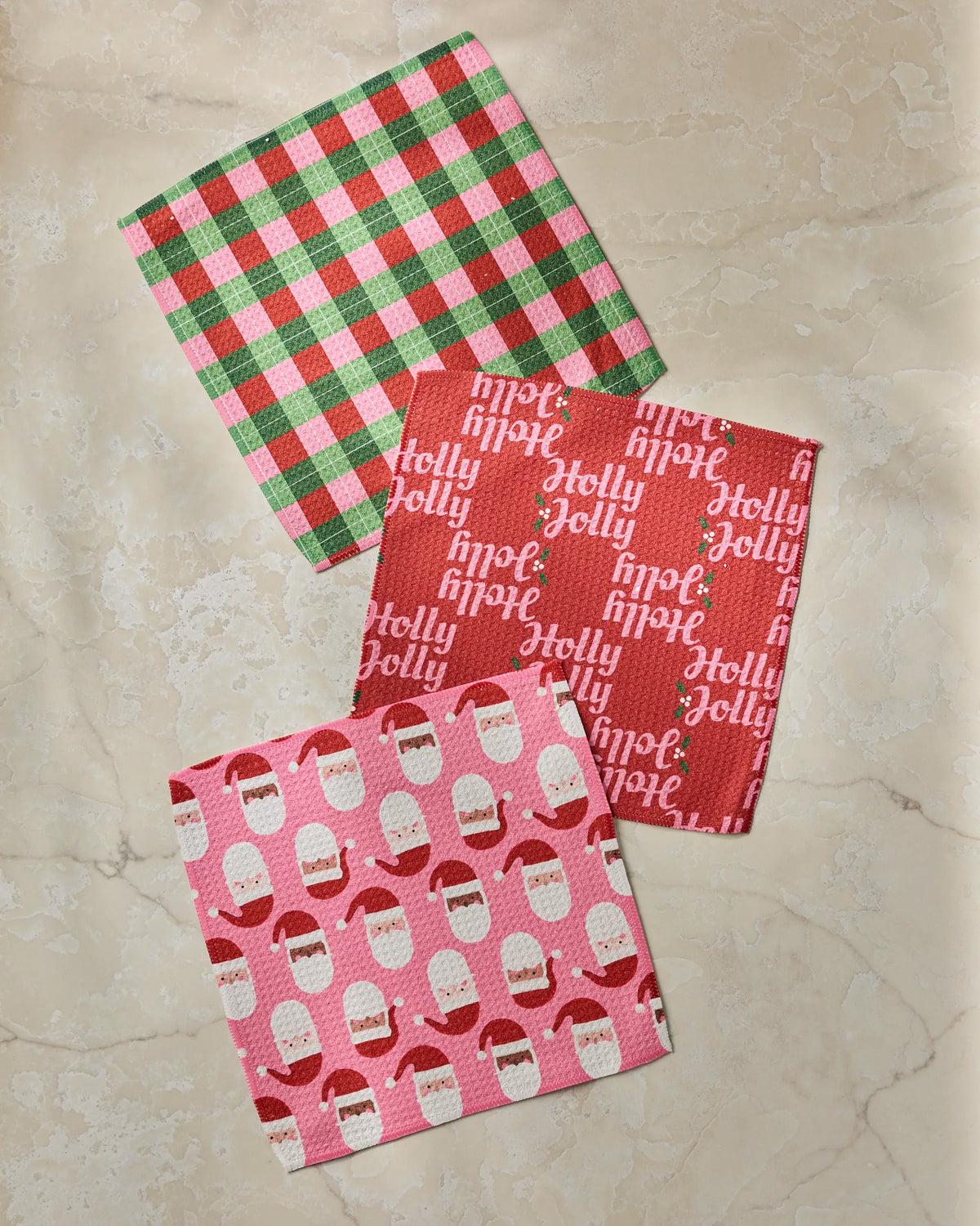Three holiday-themed napkins from Geometry’s Holly Jolly Santa collection—a green and red checkered design, a red napkin featuring “Holly Jolly” text, and a pink option adorned with white and red Santa faces—create a festive set that’s perfect for holiday gatherings. Cheerful and versatile, these napkins pair beautifully with your collection of recycled dishcloths or super-absorbent dishtowels.