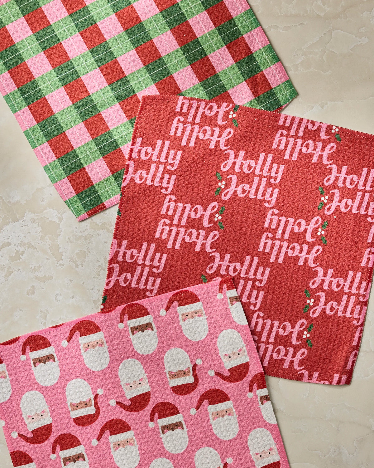 Three festive cloths from the Geometry Holly Jolly Santa set: one featuring green, red, and white plaid; one in red adorned with “Holly Jolly” and holly berries; and one pink with a pattern of Santa faces—all displayed on a light beige surface. These recycled dishcloths are both eco-friendly and stylish, making them the perfect addition to your holiday kitchen.