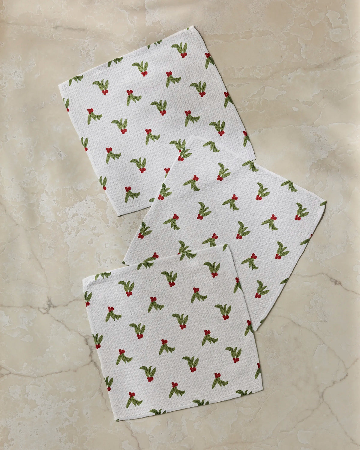 Three Geometry “Holly Sprinkle” white napkins, featuring a subtle holly and berry pattern, are arranged on a light marble surface. Reminiscent of a festive dishcloth set, the napkins overlap at the center and are placed at gentle angles.