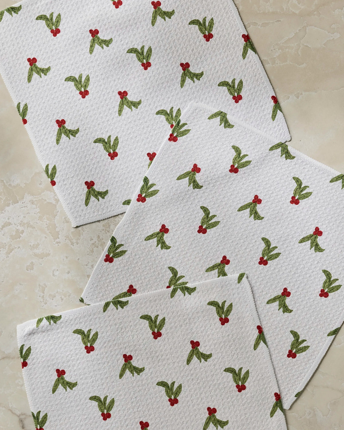 Three “Holly Sprinkle” napkins by Geometry, featuring a pattern of green holly leaves and red berries on a white background, are arranged on a light-colored marble surface for a festive accent. These absorbent kitchen towels add cheer to any dishcloth set while providing practical use for all your holiday gatherings.