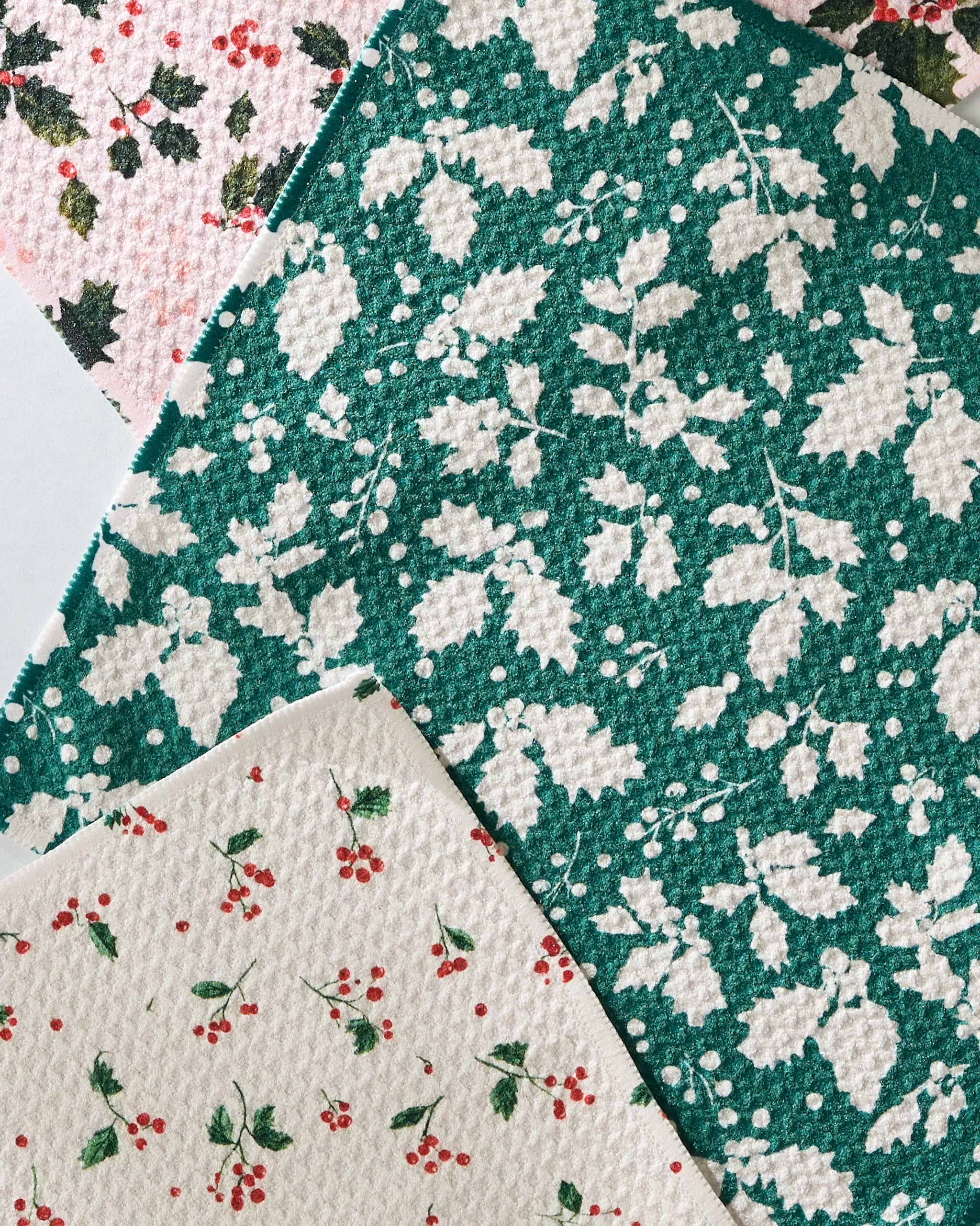 The Geometry Hollyday set features three textured, absorbent kitchen towels with festive patterns: green with white holly leaves, cream with red berries, and pink with holly leaves and berries, beautifully layered together.