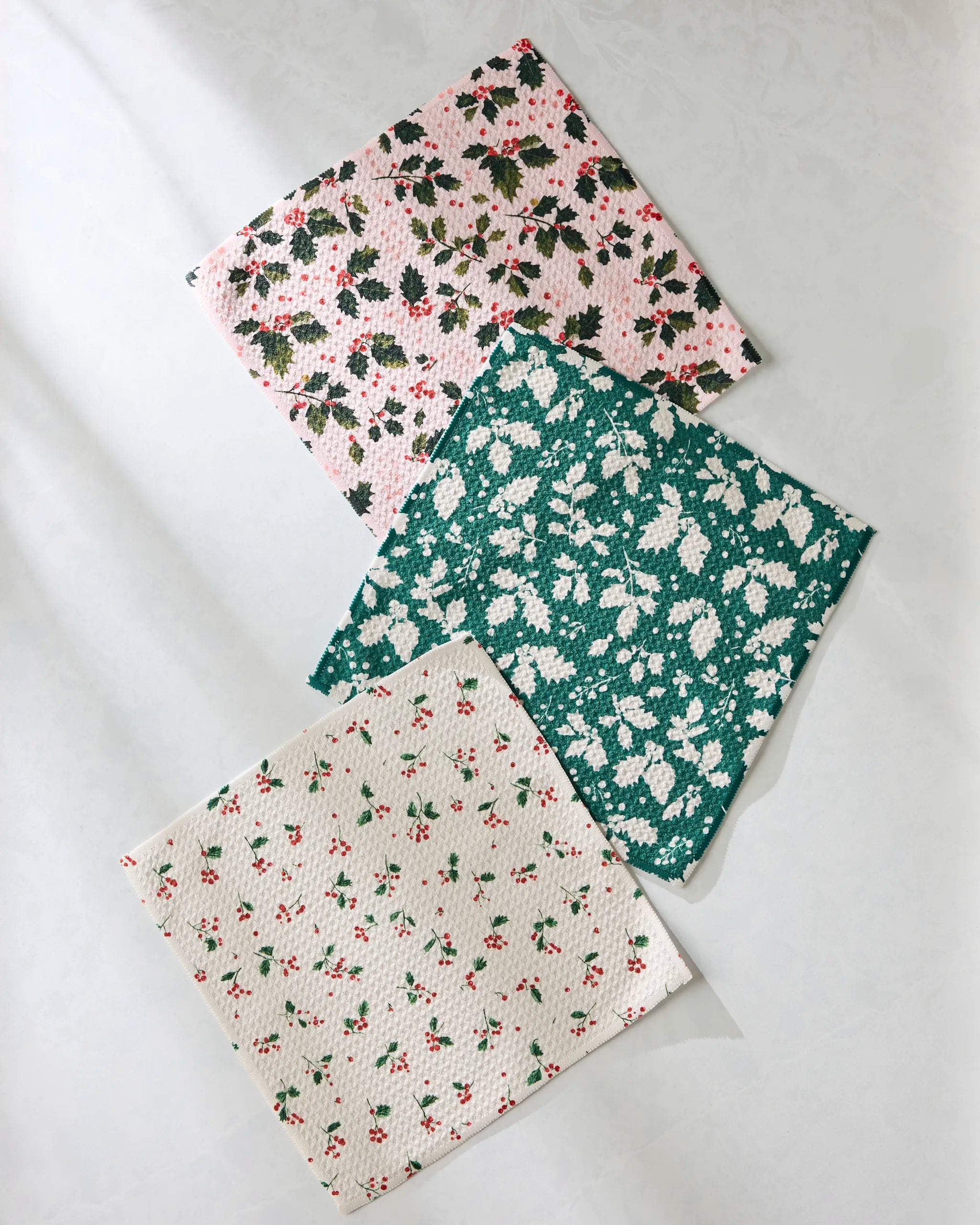 Three absorbent Geometry Hollyday kitchen towels—one pink with small flowers, one green with white floral designs, and one white with tiny red flowers—are arranged on a light surface.
