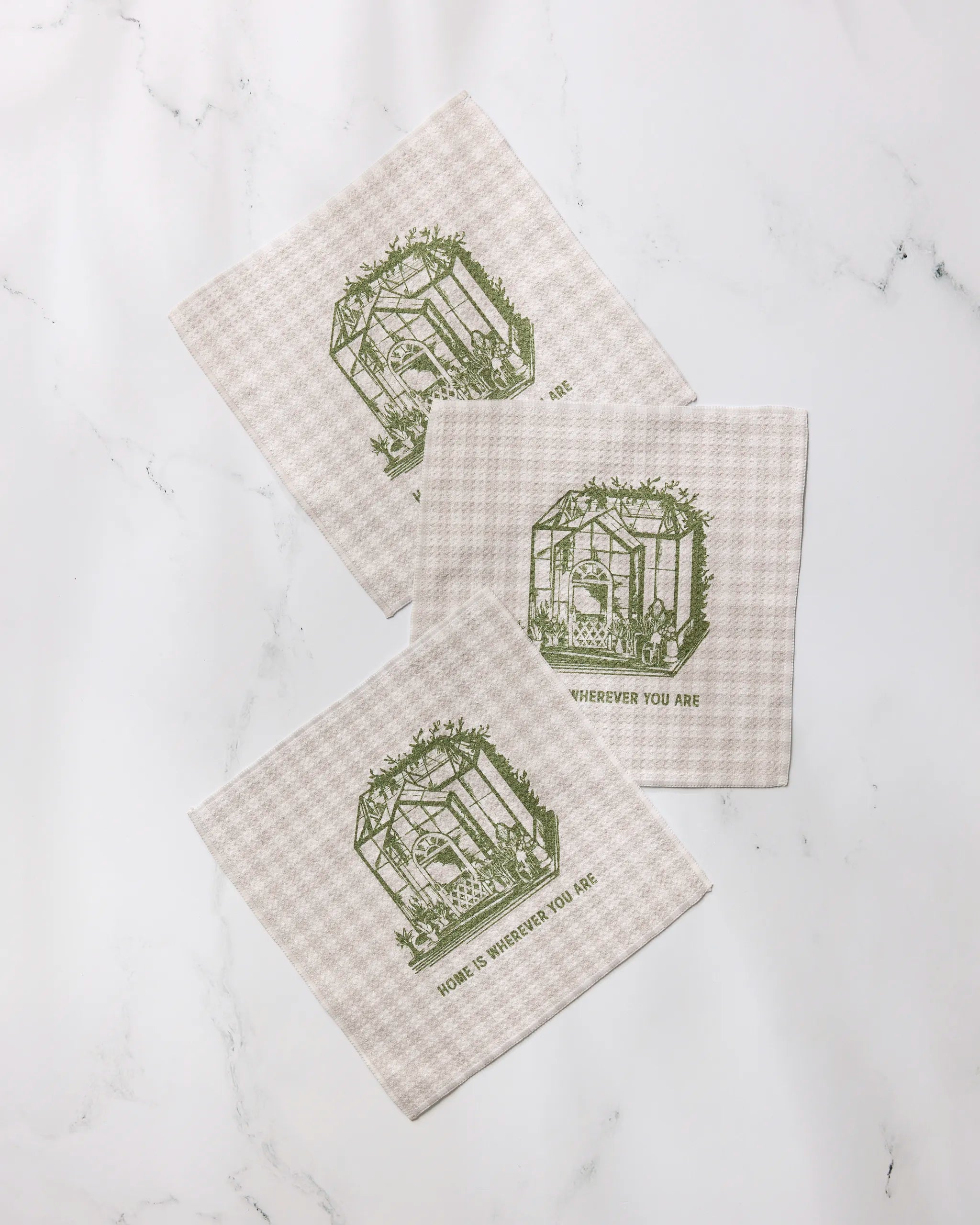 Three beige checkered recycled cloth napkins from Geometry’s “Home is You” collection, featuring green greenhouse illustrations and the phrase “Home is wherever you are,” displayed on a white marble surface.
