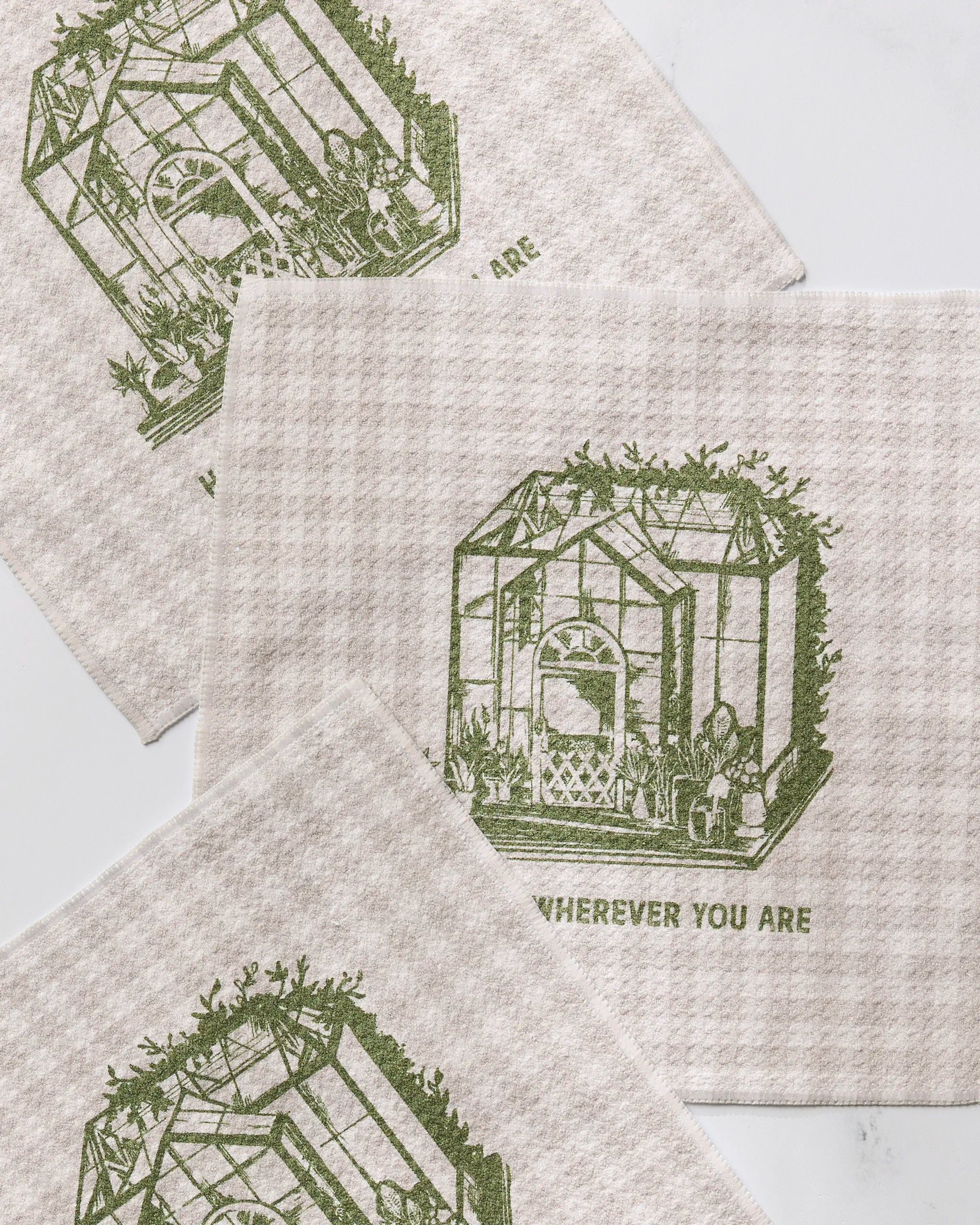 Three beige cloths from Geometry’s "Home is You" recycled kitchen set feature a green greenhouse illustration and the words “WHEREVER YOU ARE,” arranged slightly overlapping on a light background.
