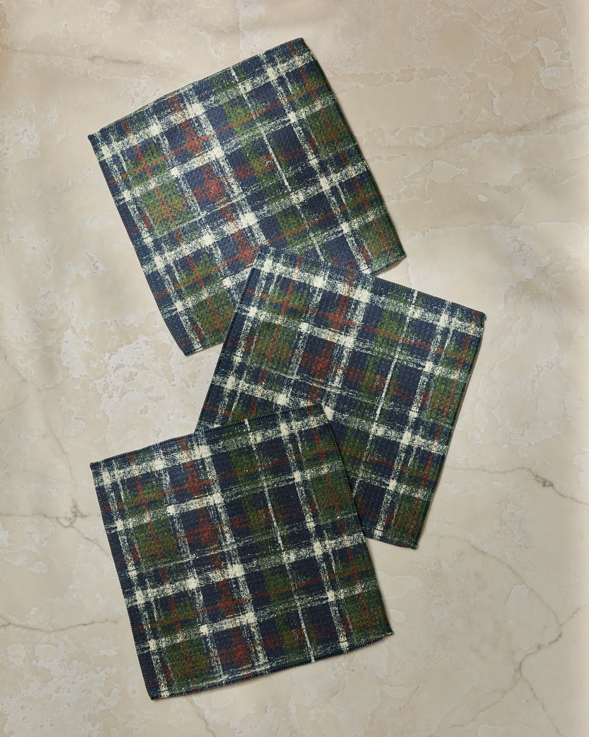 Three folded Homestead Plaid cloth napkins by Geometry, featuring a blue, green, red, and white checkered pattern and crafted from recycled materials, are arranged on a light beige marble surface.