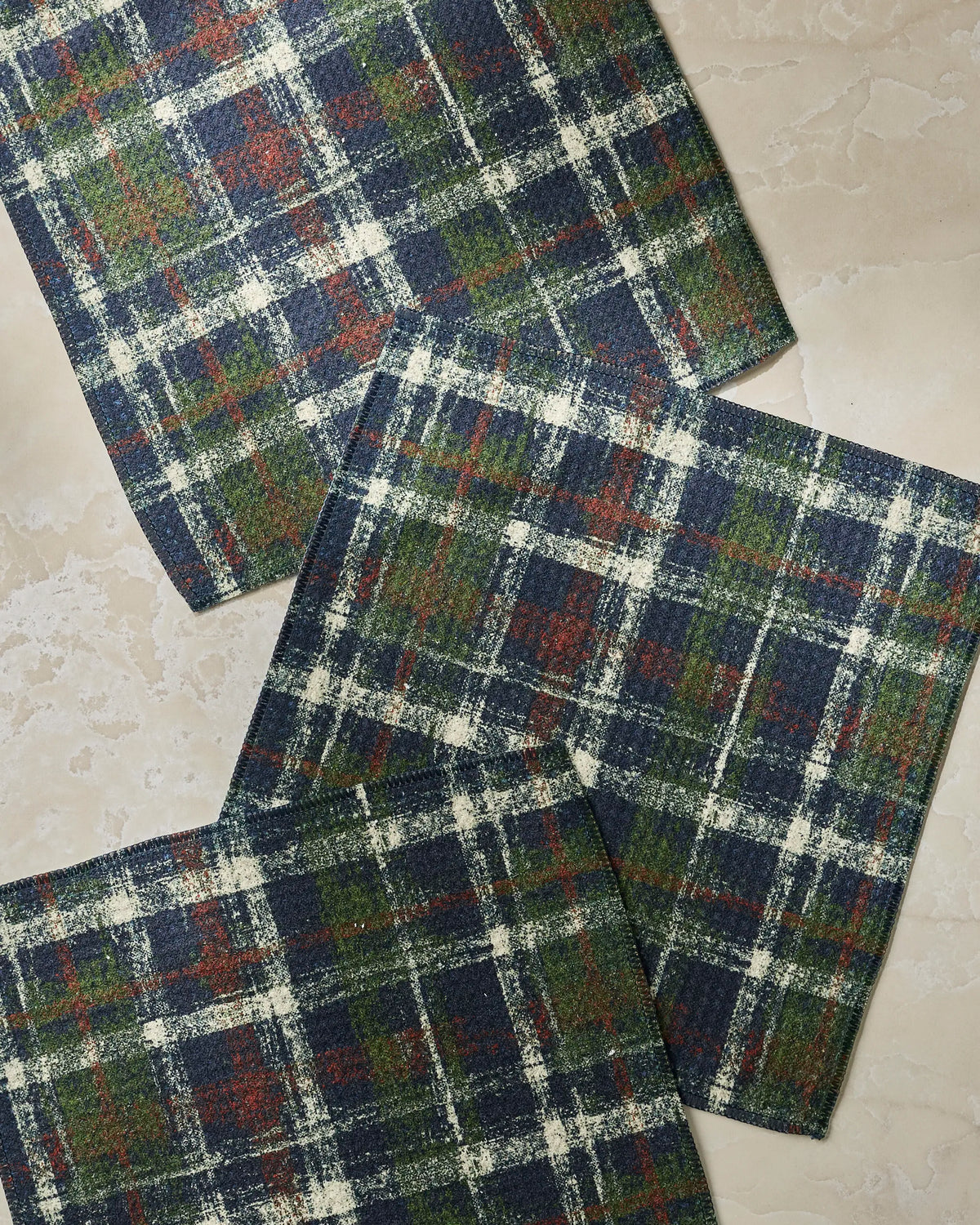 Three rectangular pieces of fabric featuring the Homestead Plaid pattern by Geometry—showcasing navy, green, white, and red hues—are arranged on a light beige, marble-like surface. This set of super-absorbent dishtowels is thoughtfully crafted from recycled materials and adds a classic touch to your kitchen.