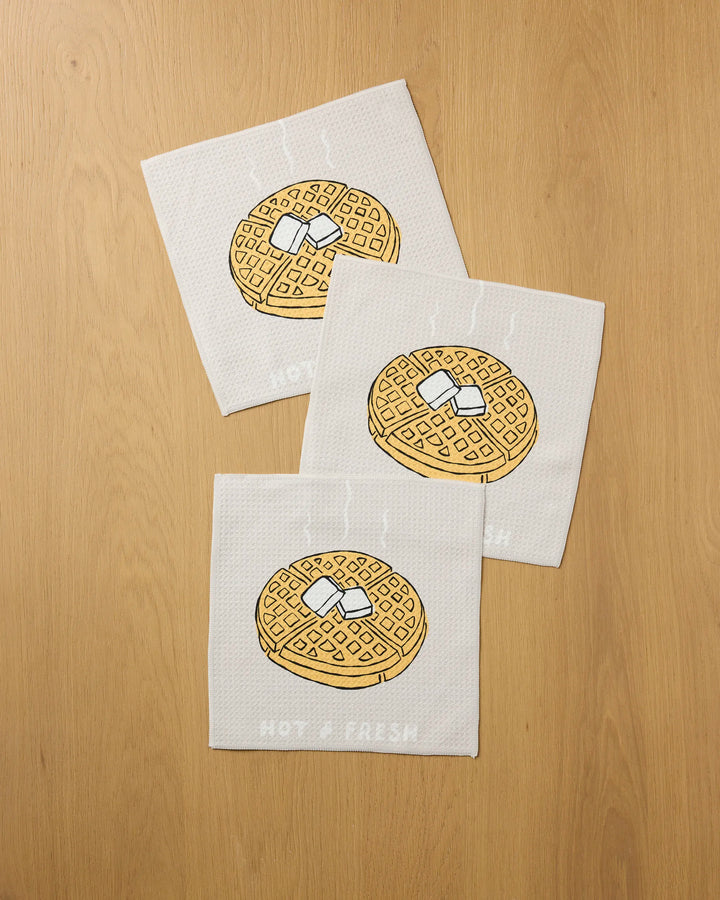 Three white "Hot and Fresh" dish towels by Rosa Kichline, featuring steaming waffle illustrations with butter and syrup and the words HOT & FRESH below each design, are displayed on a wooden surface.