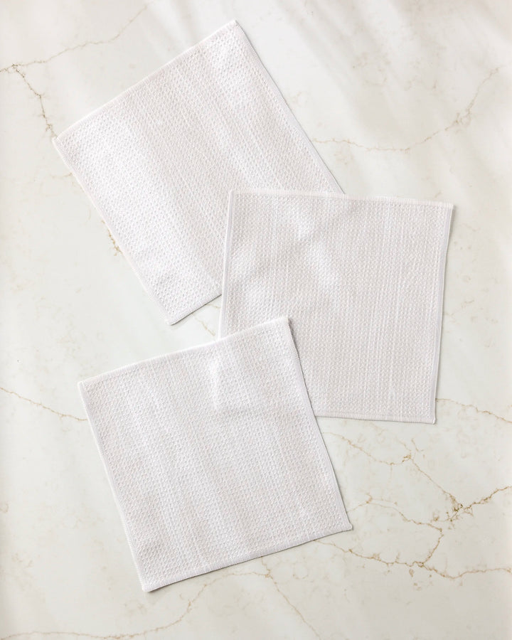 Three white, textured Houston Wood dishcloths by Kailee Wright, made from recycled materials, are displayed on a light marble surface with subtle veins. The set overlaps slightly and is arranged at varying angles.