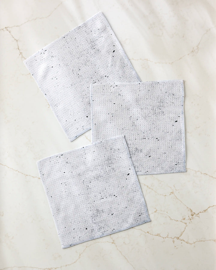 Three grey, textured Hudson Grey dishcloths by Kailee Wright—part of a super-absorbent dishtowel set—are arranged diagonally on a light marble surface with beige veins.