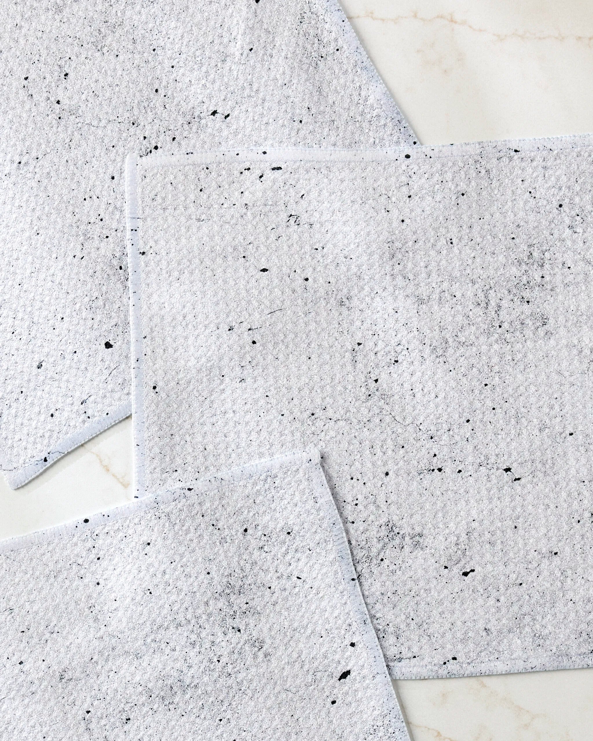 Three Hudson Grey cloths by Kailee Wright, made from recycled materials, feature a white textured design with black speckles and are displayed overlapping on a light marble surface.