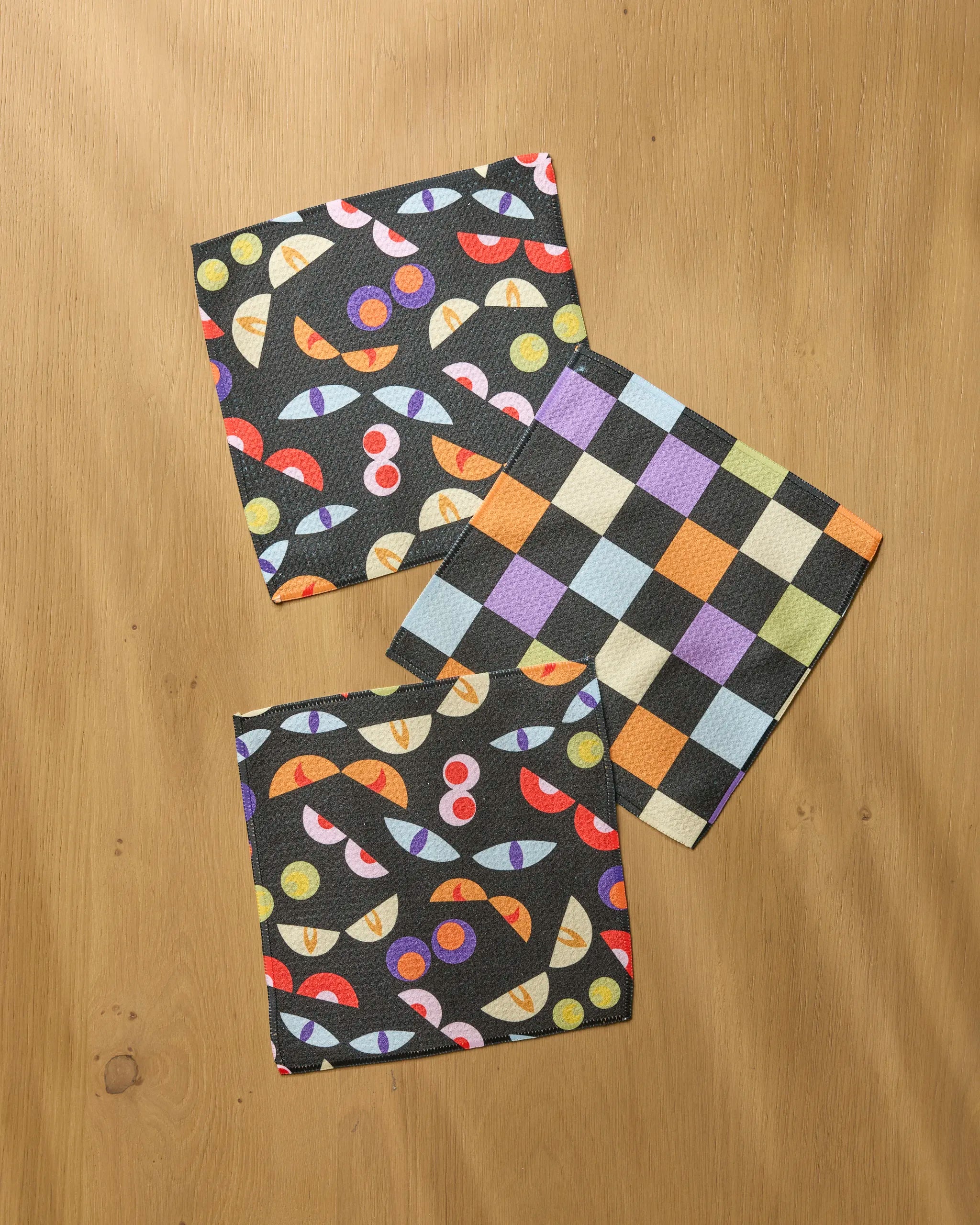 Add flair to your kitchen with Geometry’s "I See You"—three folded cloth napkins on wood: two have colorful abstract designs, one has a pastel checkered pattern. Perfect as absorbent dish towels or to brighten your dishcloth set.