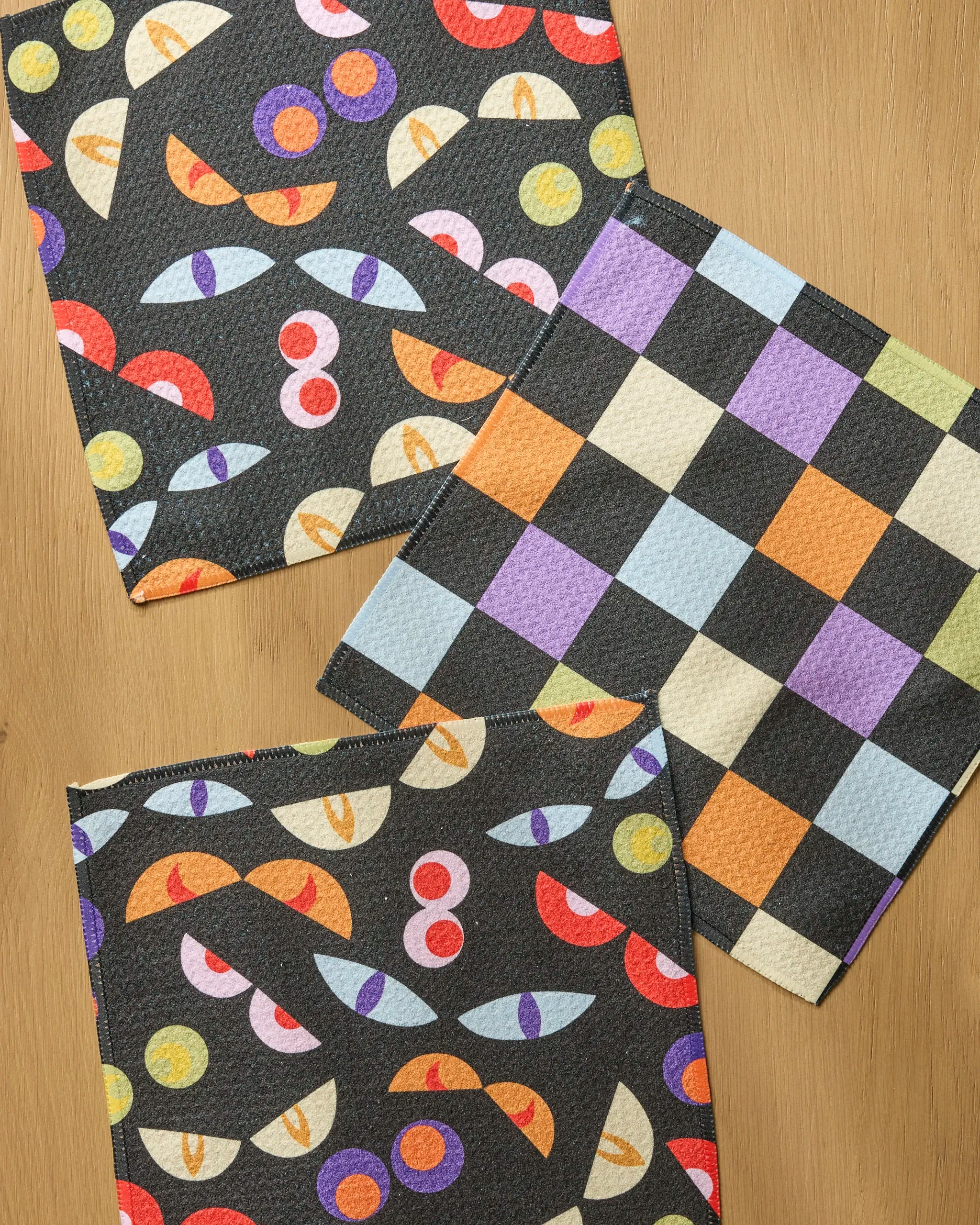 Three "I See You" recycled dishcloths by Geometry rest on wood: two show abstract eyes and circles in bold colors on black; one features a pastel checkerboard in orange, green, purple, black, and white.
