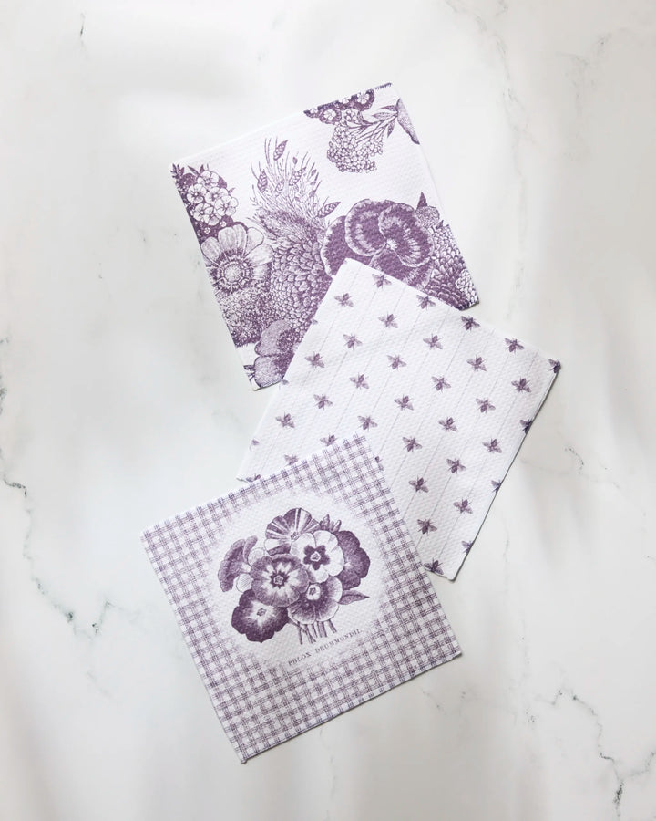 Three folded Ikebana dish towels by Geometry—a set with floral and fruit prints, bees, and a floral check design—are arranged on a white marble surface.