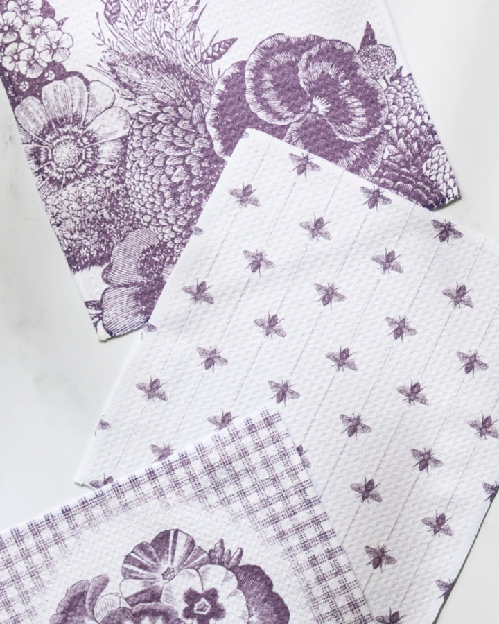 The Ikebana dishcloth set by Geometry includes three absorbent, white cloths with purple floral and bee designs plus a checkered pattern with a floral border—combining style and function for your kitchen.