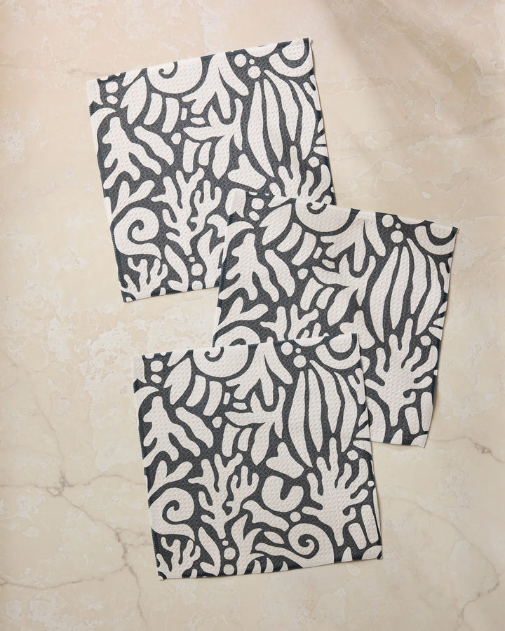Three "Inkwell Reef" square paper napkins by Geometry, featuring a black and white abstract floral and leaf pattern, are overlapped on a light marble surface. Made from recycled materials.