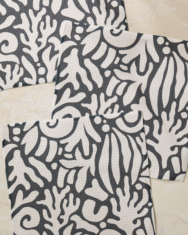 Three Geometry "Inkwell Reef" paper napkins, made from recycled materials, display a bold abstract black and white botanical pattern and are arranged slightly overlapping on a light marble-like surface.
