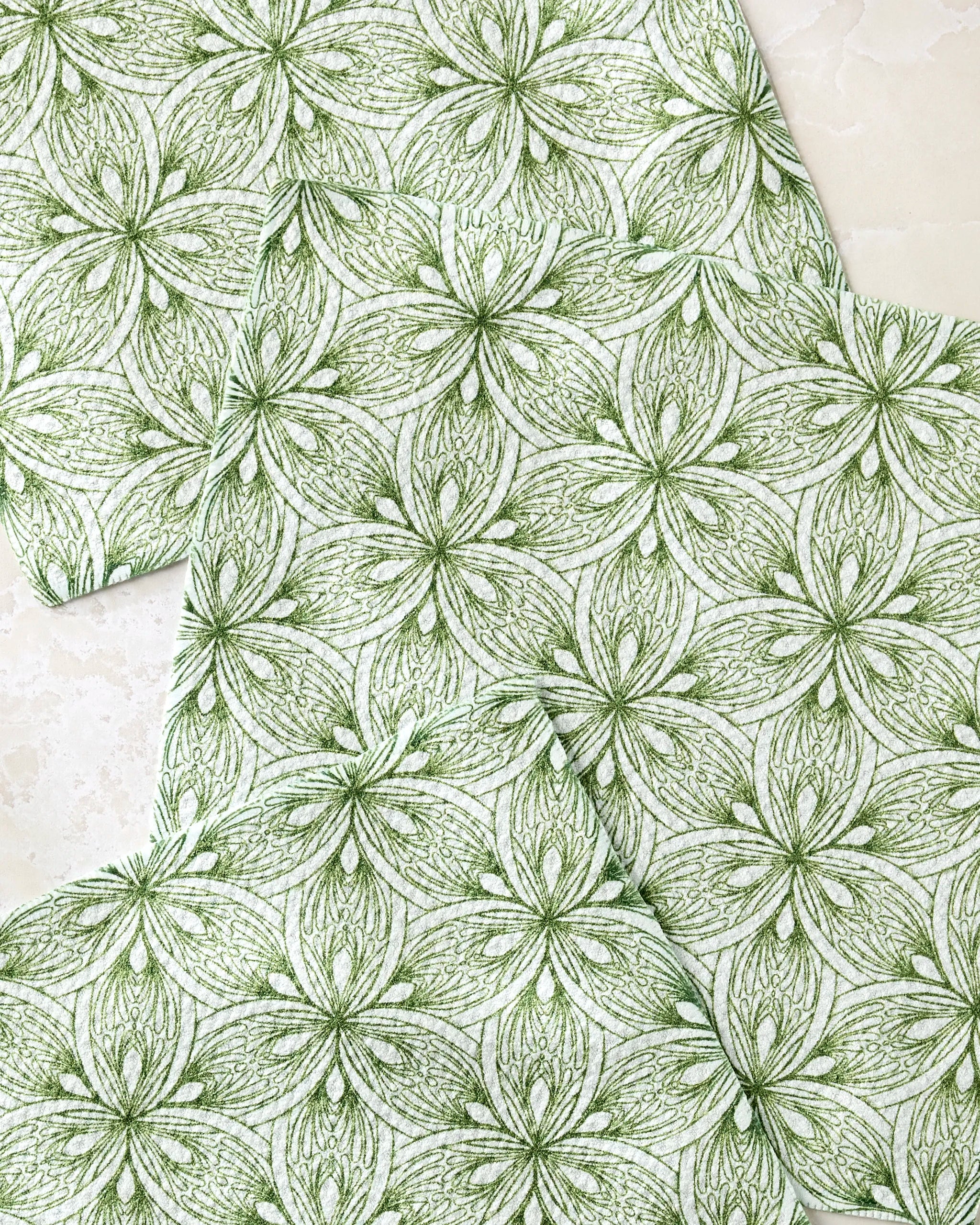 Three "Interlaced Petals" square napkins by Marta Harvey, made from recycled materials, display a green-and-white floral and leaf pattern. The napkins are arranged overlapping on a light textured surface.