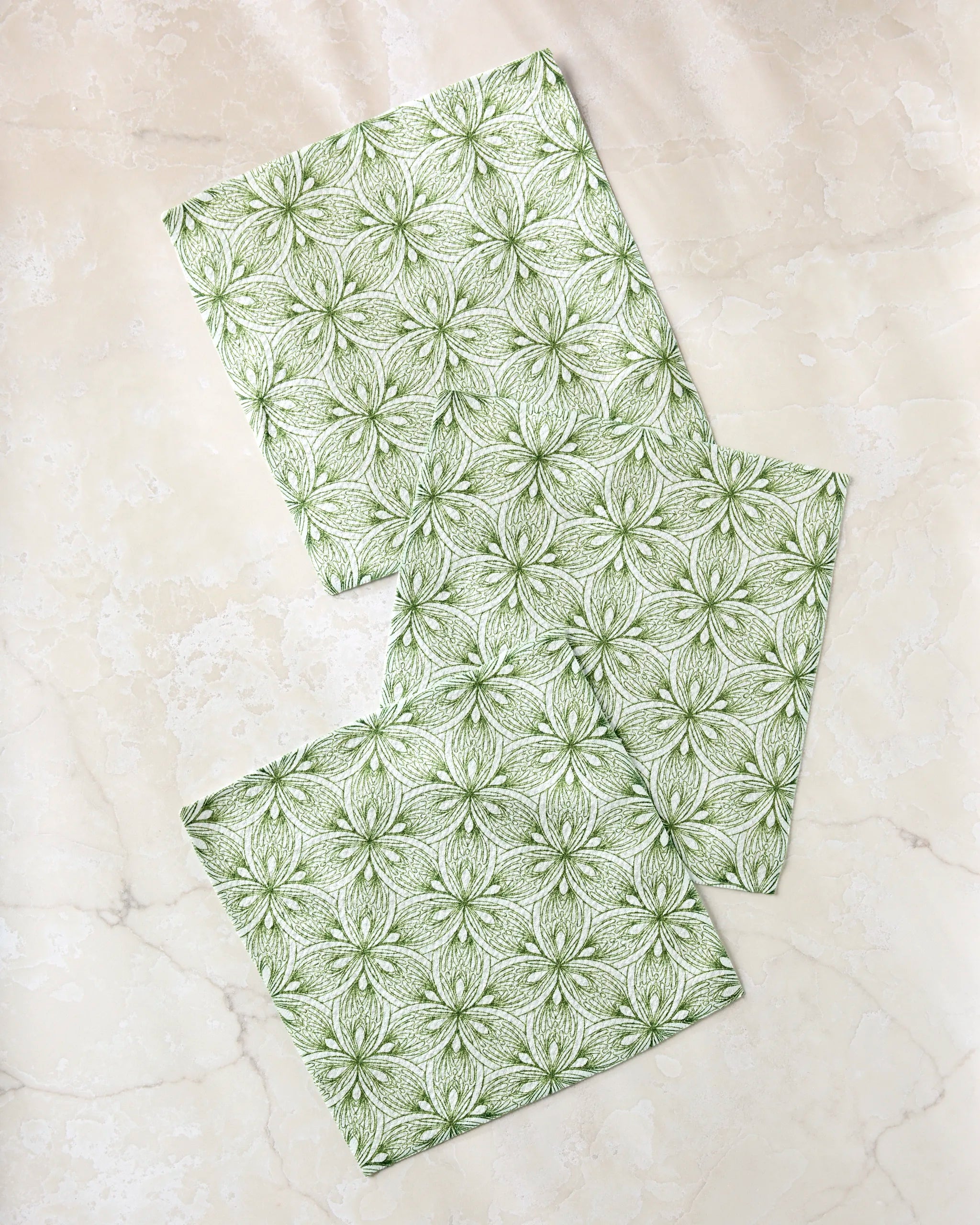Three green and white Interlaced Petals cloth napkins by Marta Harvey, with a floral geometric design, are part of a super-absorbent dishtowel set and arranged overlapping on a light cream marble surface.