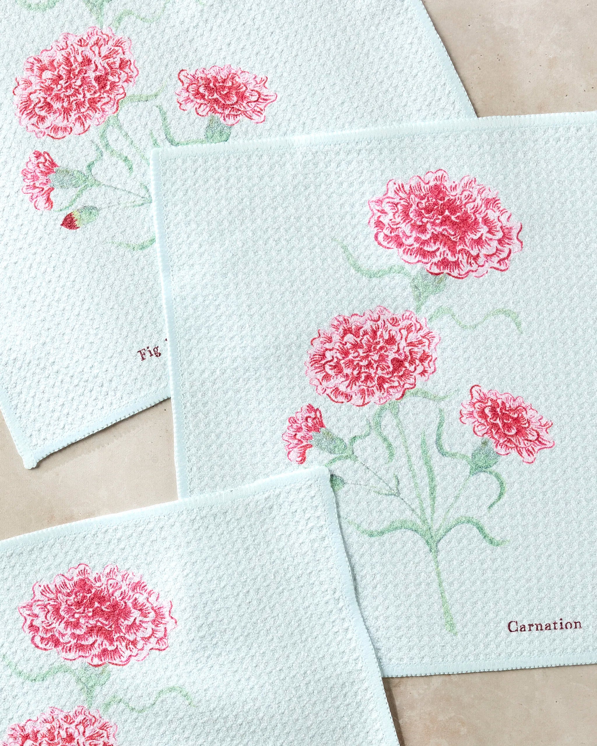 Three white textured cloths from Linda Fox’s January Carnation line, made with recycled materials, feature pink carnation designs; one says “Carnation,” another is marked “1/1.” They are displayed on a light-colored surface.