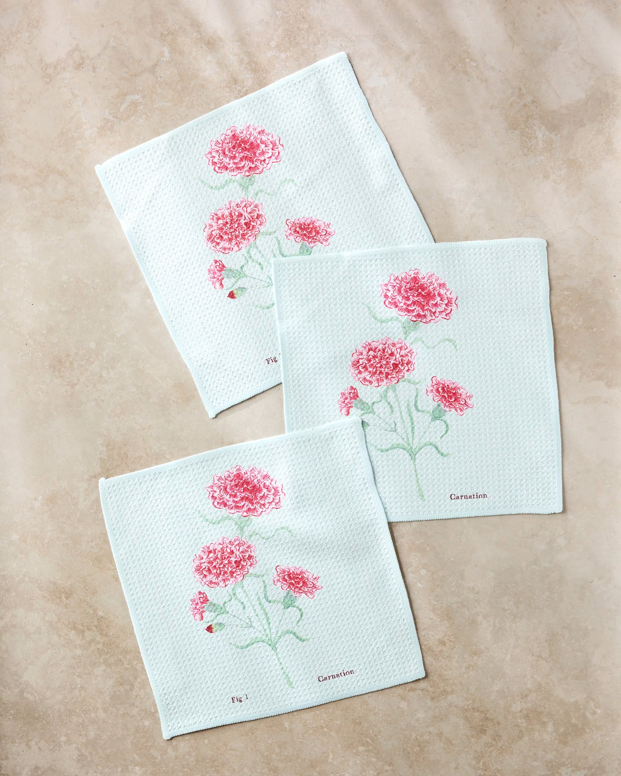 Three January Carnation cloth napkins by Linda Fox, crafted from recycled light blue fabric and embroidered with pink carnations and green stems, are neatly displayed on a beige marble surface.