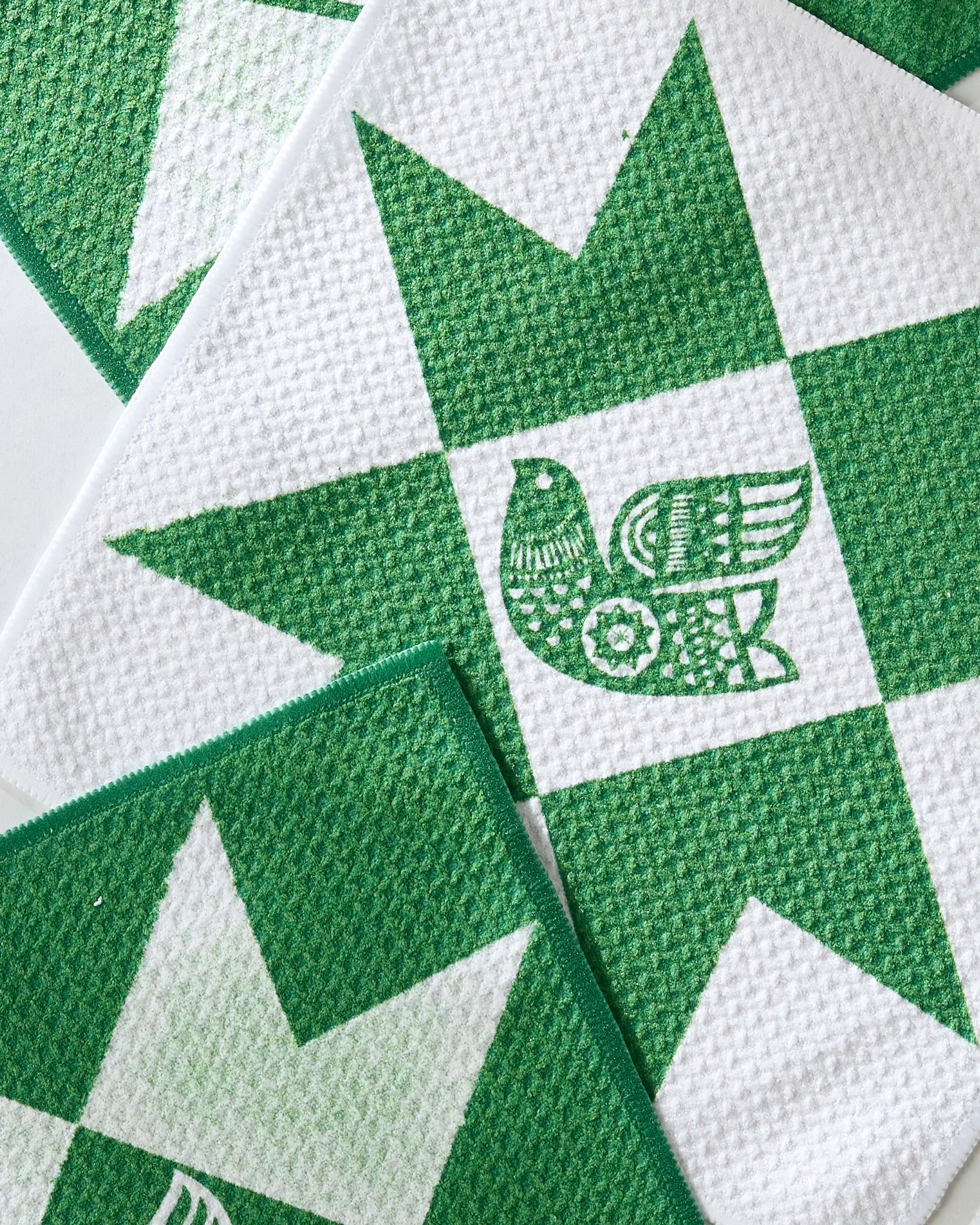 The Joybird reusable dishcloth set by Geometry features white textured cloths with green geometric star patterns, each with a stylized bird at the center—overlapping in the image, ideal for an eco-friendly kitchen.