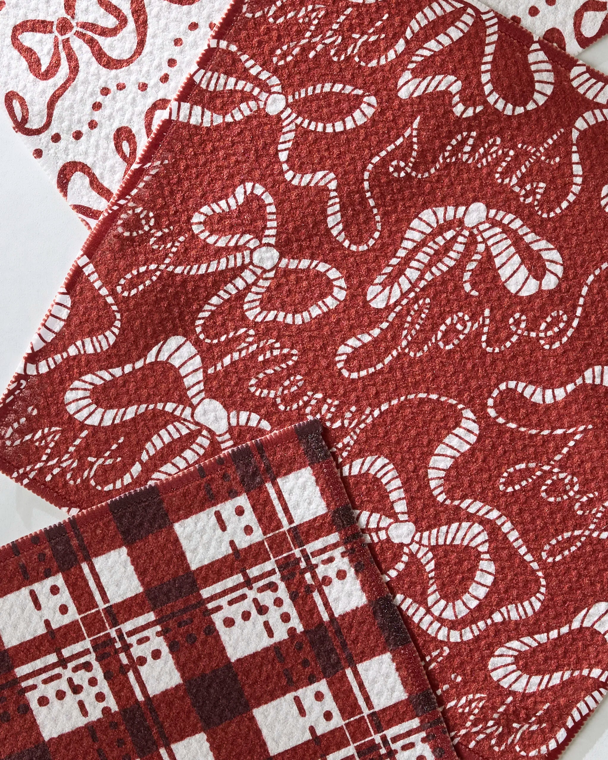 Three "Joyful Night" dishcloths by Geometry, made from recycled materials, display festive bow and ribbon, plaid checkered, and swirl designs, and are arranged overlapping on a plain surface.