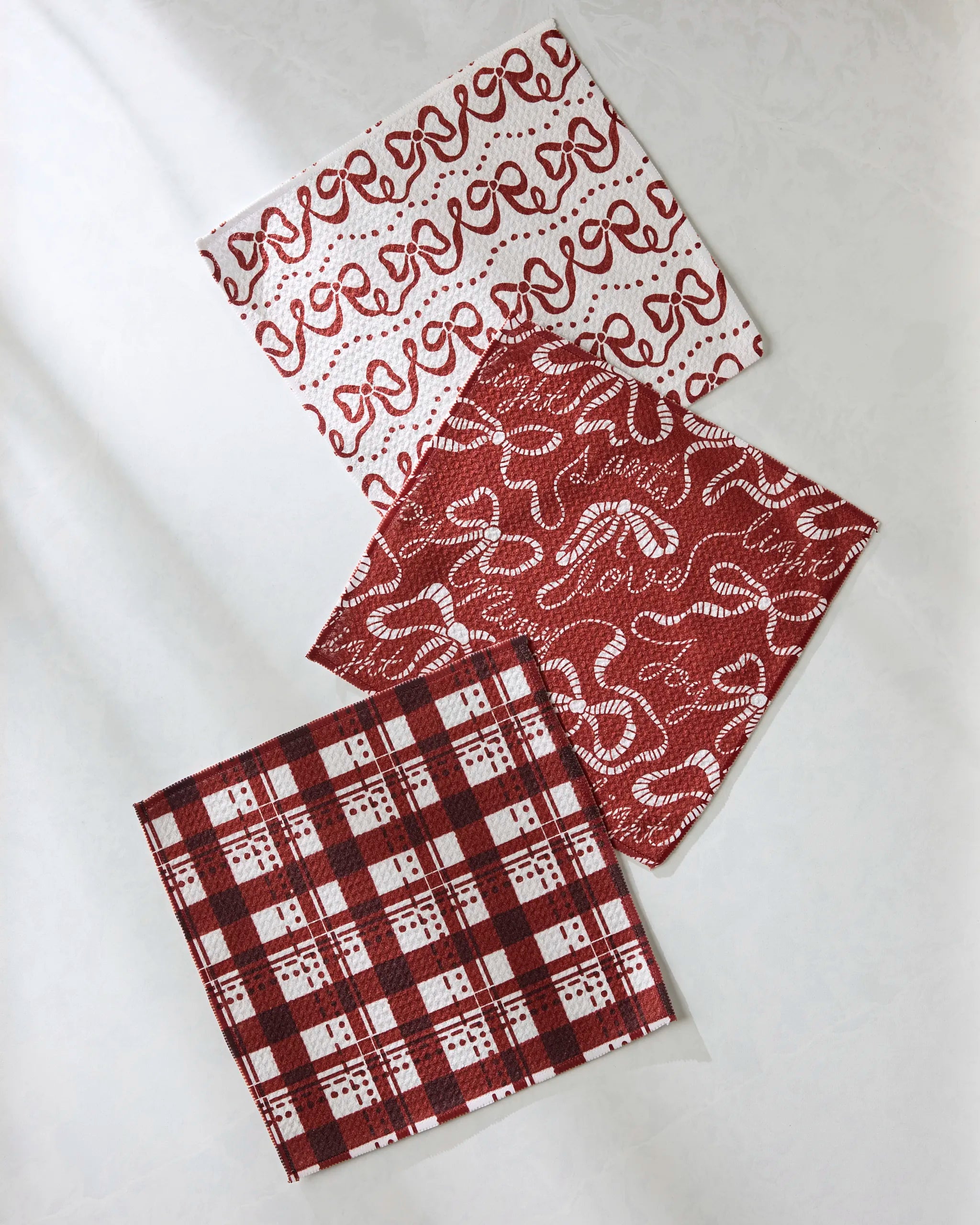 Three folded Joyful Night dishcloths by Geometry rest on a white surface: one with cursive "love" and hearts, one with swirls and hearts, and one with a red and white plaid checkered design.