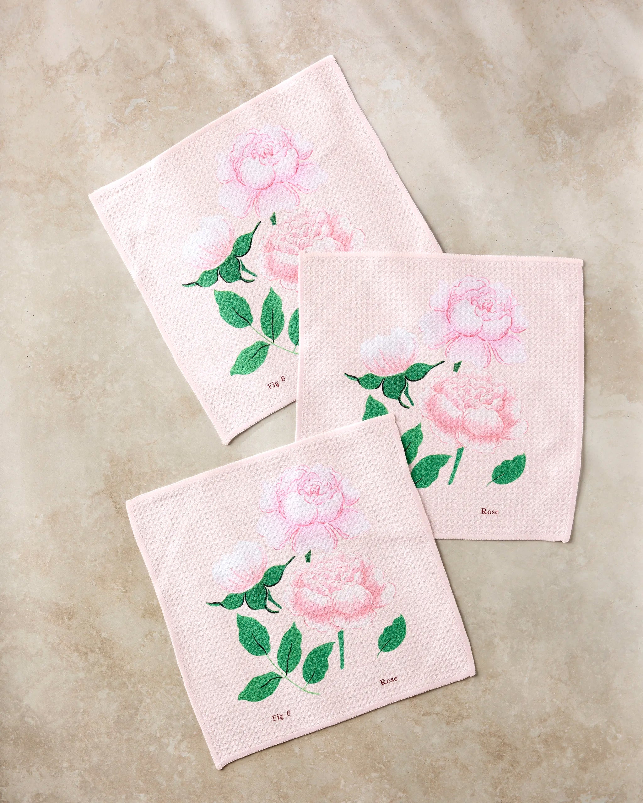 Three pale pink June Roses dishcloths by Linda Fox, embroidered with light pink peonies and green leaves, are arranged on a beige marble surface. Each features "Rosa" and "Tea" labeled near the floral designs.