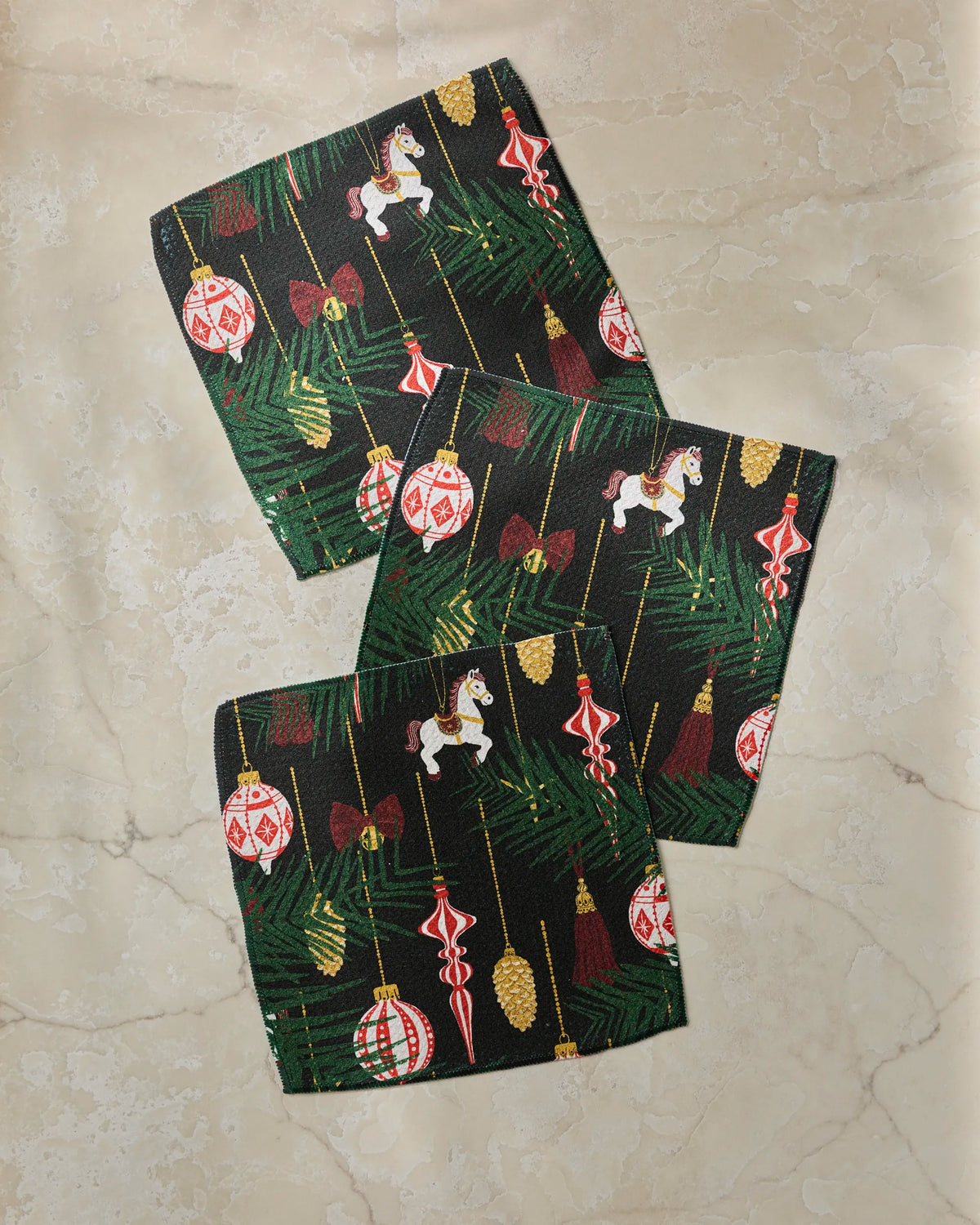 Three fabric squares from Geometry’s “Keepsake” collection, showcasing a festive Christmas pattern with ornaments, rocking horses, pine branches, and bows on a marble surface—perfect as absorbent kitchen towels or as part of your recycled dishcloth set for the holiday season.
