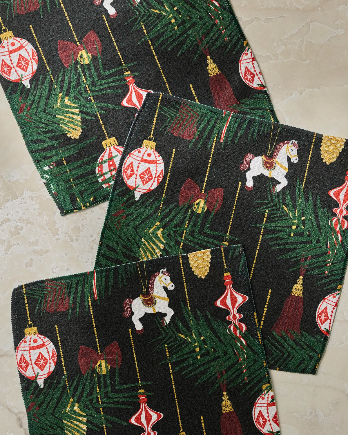 Three rectangular black Keepsake placemats by Geometry showcase festive Christmas designs—including pine branches, ornaments, bows, pinecones, and toy horses—and are arranged on a beige marble surface. For a perfectly coordinated and convenient holiday table setting, pair these placemats with an absorbent set of Geometry kitchen towels.