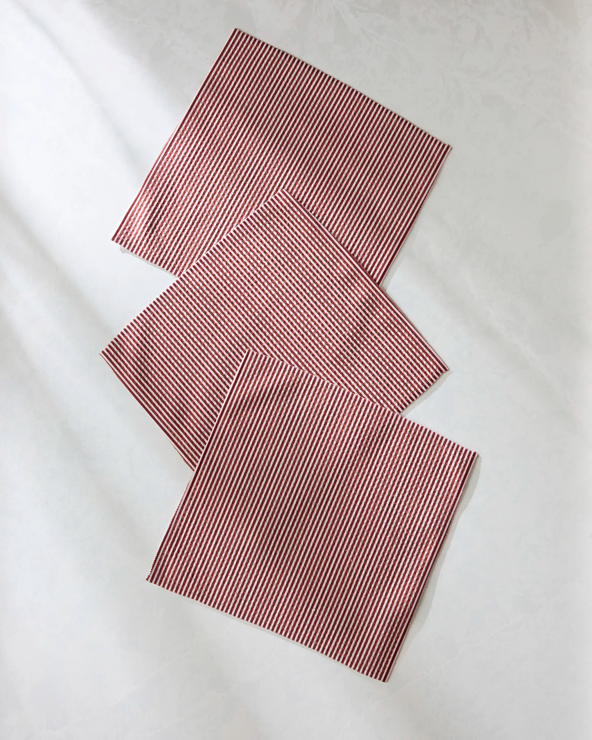 Three Geometry Kringle napkins, featuring red and white stripes and made from recycled materials, are neatly arranged in a diagonal line on a plain surface, creating an eye-catching, even pattern.