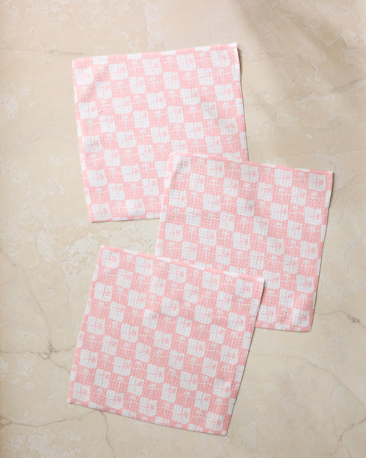 Three La Brisa Pink cloth napkins by Geometry are neatly arranged on a light beige marble surface. The napkins, with a soft, textured look and pink-and-white checkered design, overlap slightly to showcase their stylish pattern.