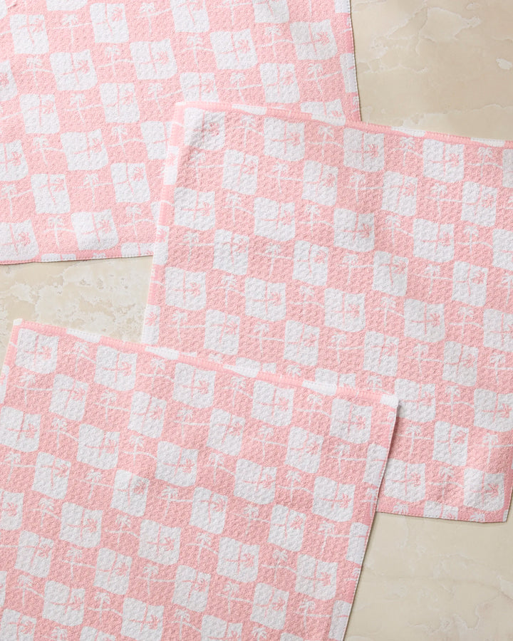 Three La Brisa Pink dish towels from Geometry, featuring pink and white checkered squares, are laid out overlapping on a light beige marble surface.
