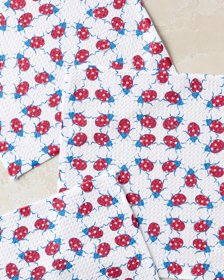 Four white "Ladybug Dance" kitchen towels by Second Nature Studio, featuring a red and blue ladybug pattern, are spread on a light stone surface with overlapping edges.