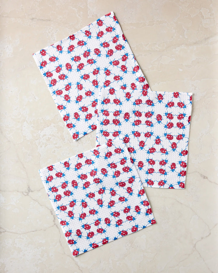 Three 'Ladybug Dance' cloth napkins by Second Nature Studio, featuring a red and blue floral pattern, are neatly folded and overlap slightly on a light beige marble surface.