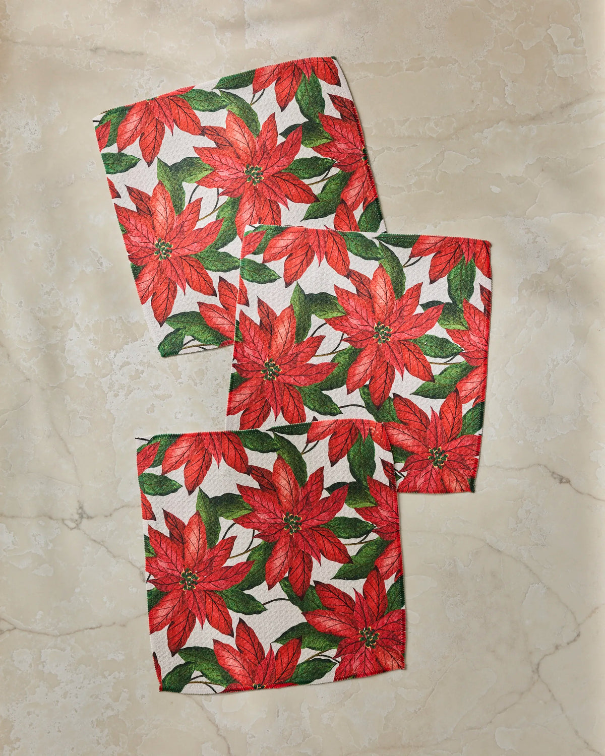 Three Lauri square napkins by Geometry, made from recycled materials, feature a festive red poinsettia and green leaf pattern, arranged diagonally on a light marbled surface.