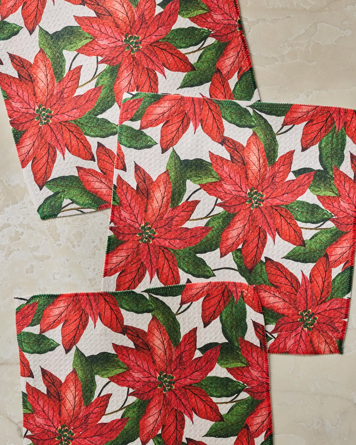 Four Lauri paper napkins by Geometry, featuring a red poinsettia and green leaf pattern, are arranged on a light beige, textured surface to create a festive accent that complements any dishcloth set.