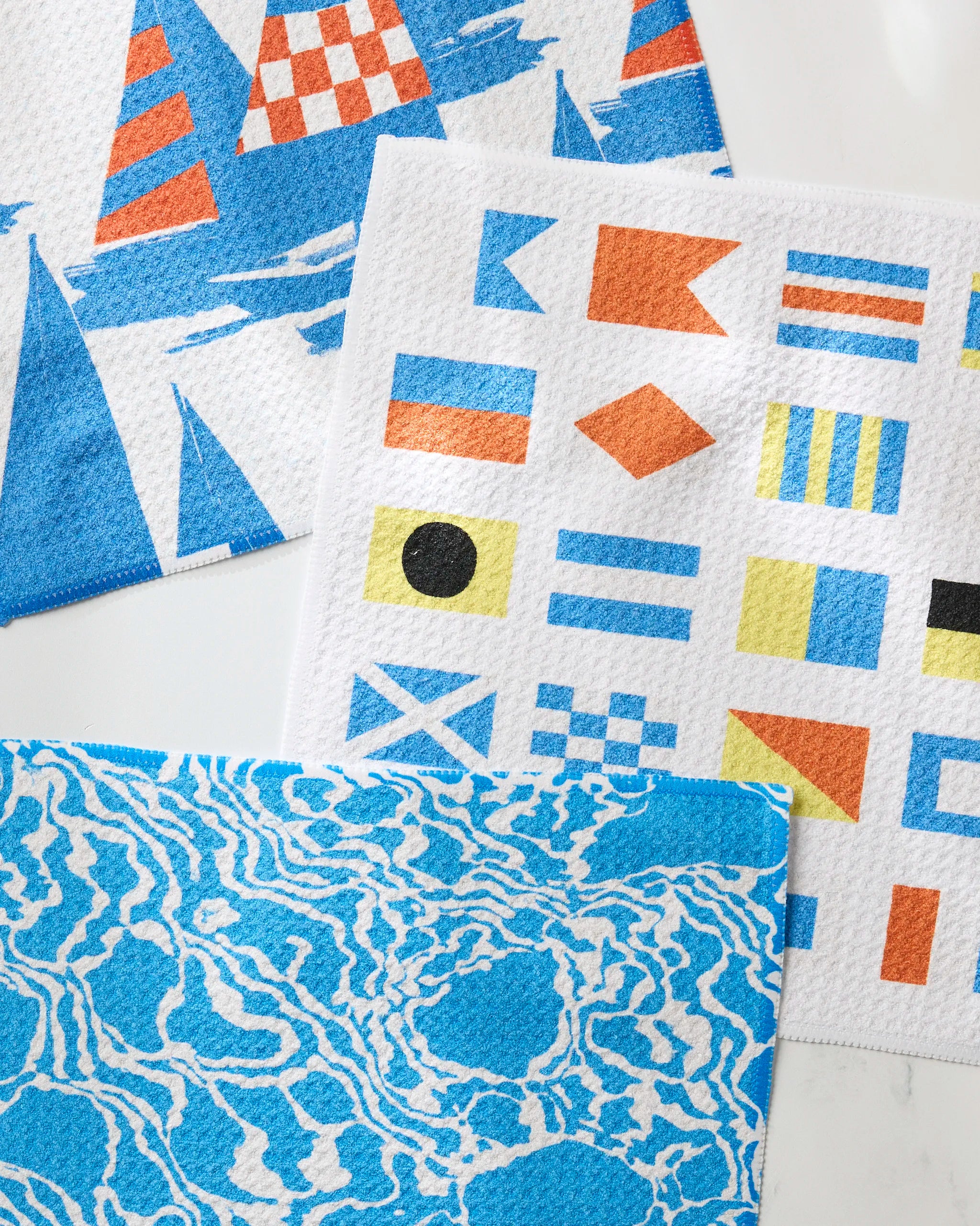 The Geometry Leeward set features three eco-friendly dishcloths with nautical designs: blue sailboats, colorful signal flags, and a blue-and-white wavy water pattern—perfect for brightening any kitchen.