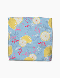 A folded blue kitchen towel from Ashley Celenas Lemon Pop set, featuring whole and sliced yellow lemons, white flowers, and pink leafy accents on super-absorbent dishcloths.