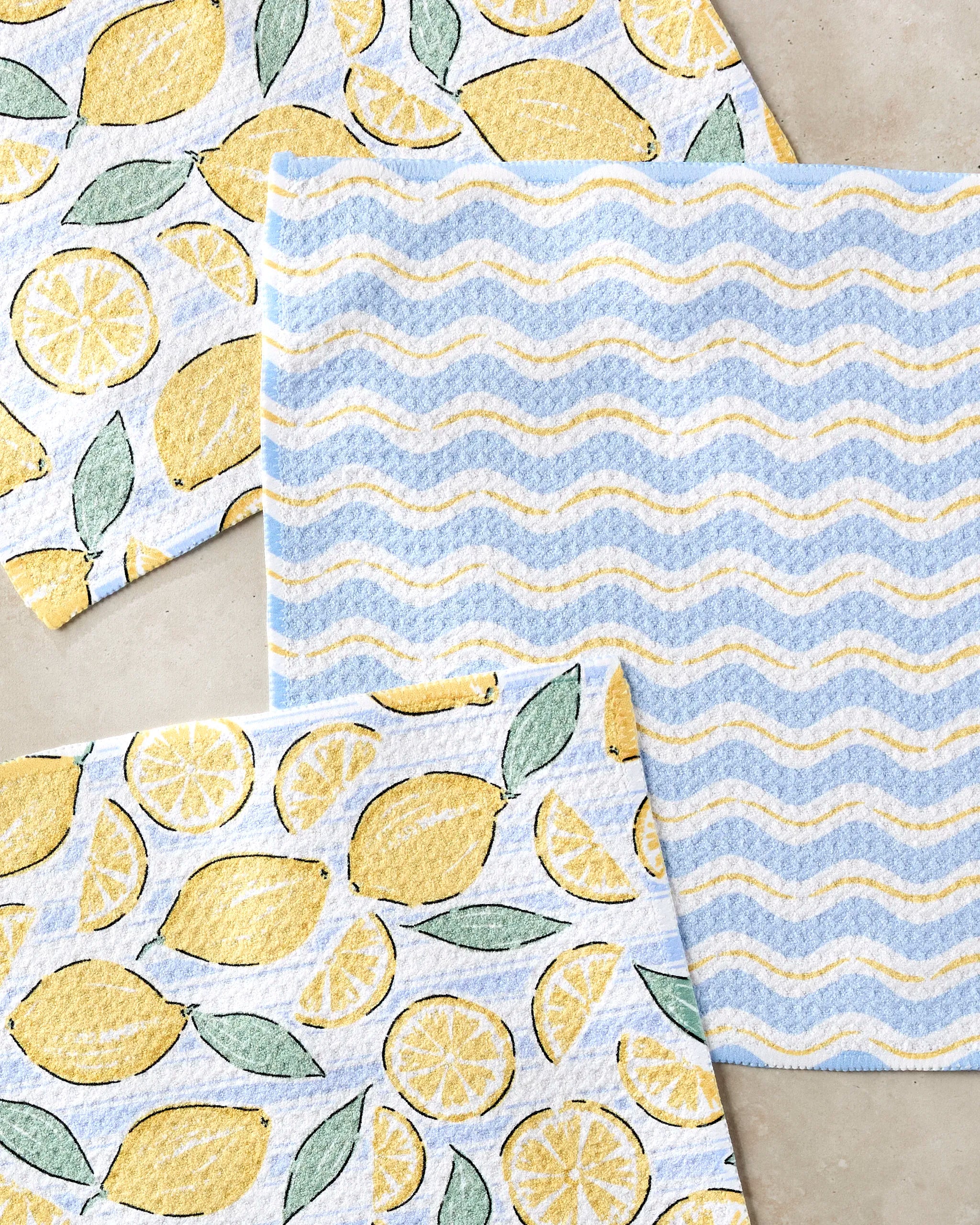 Three rectangular cloths from Hufton Studio’s Lemon Waves set are displayed: two with yellow lemons and green leaves, and one with wavy blue, white, and yellow lines, all arranged on a light surface.