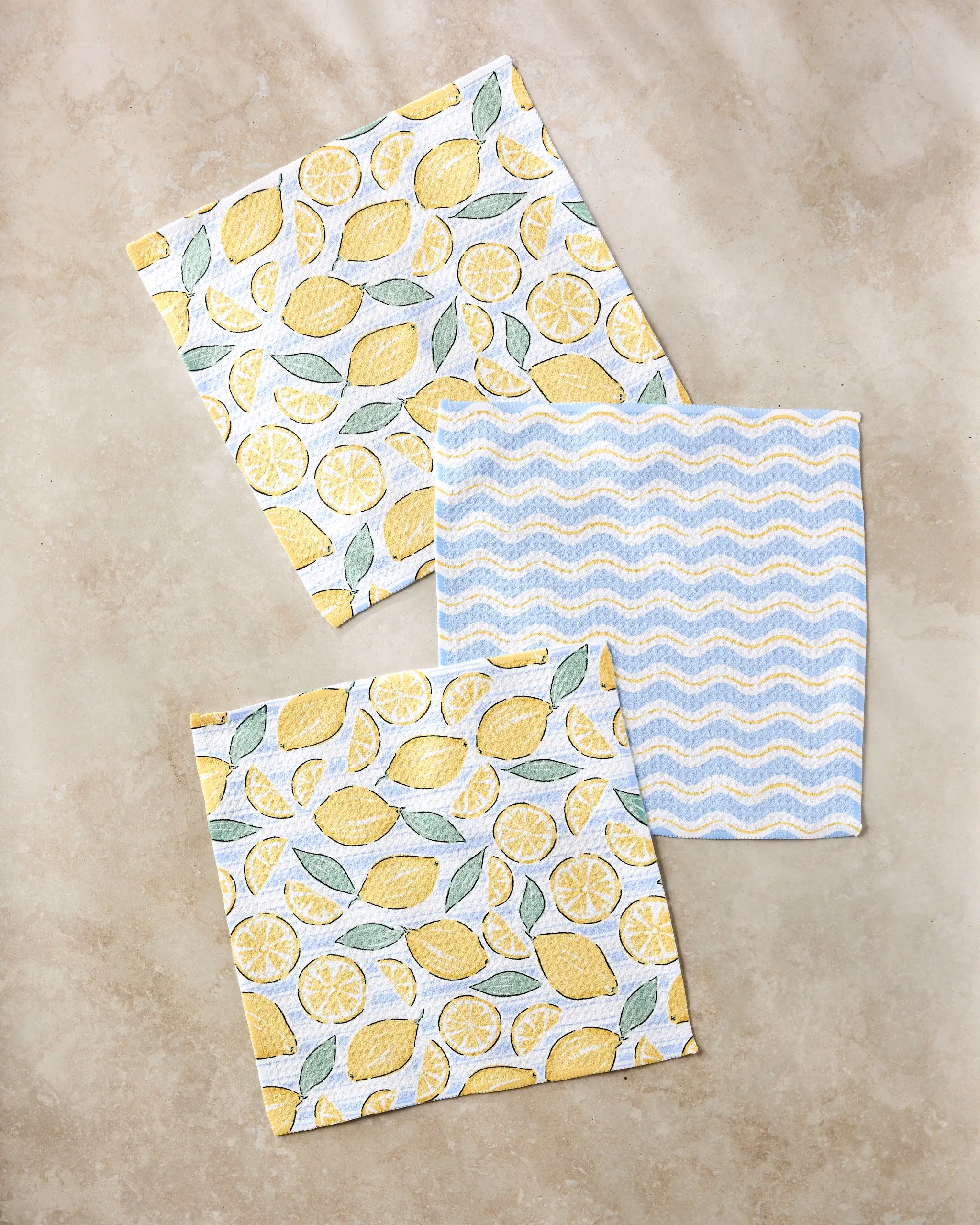 Three square Lemon Waves dishcloths by Hufton Studio are shown on a beige surface: two with a lemon and leaf pattern, and one with a blue and white zigzag design—bringing both practicality and style to your kitchen.