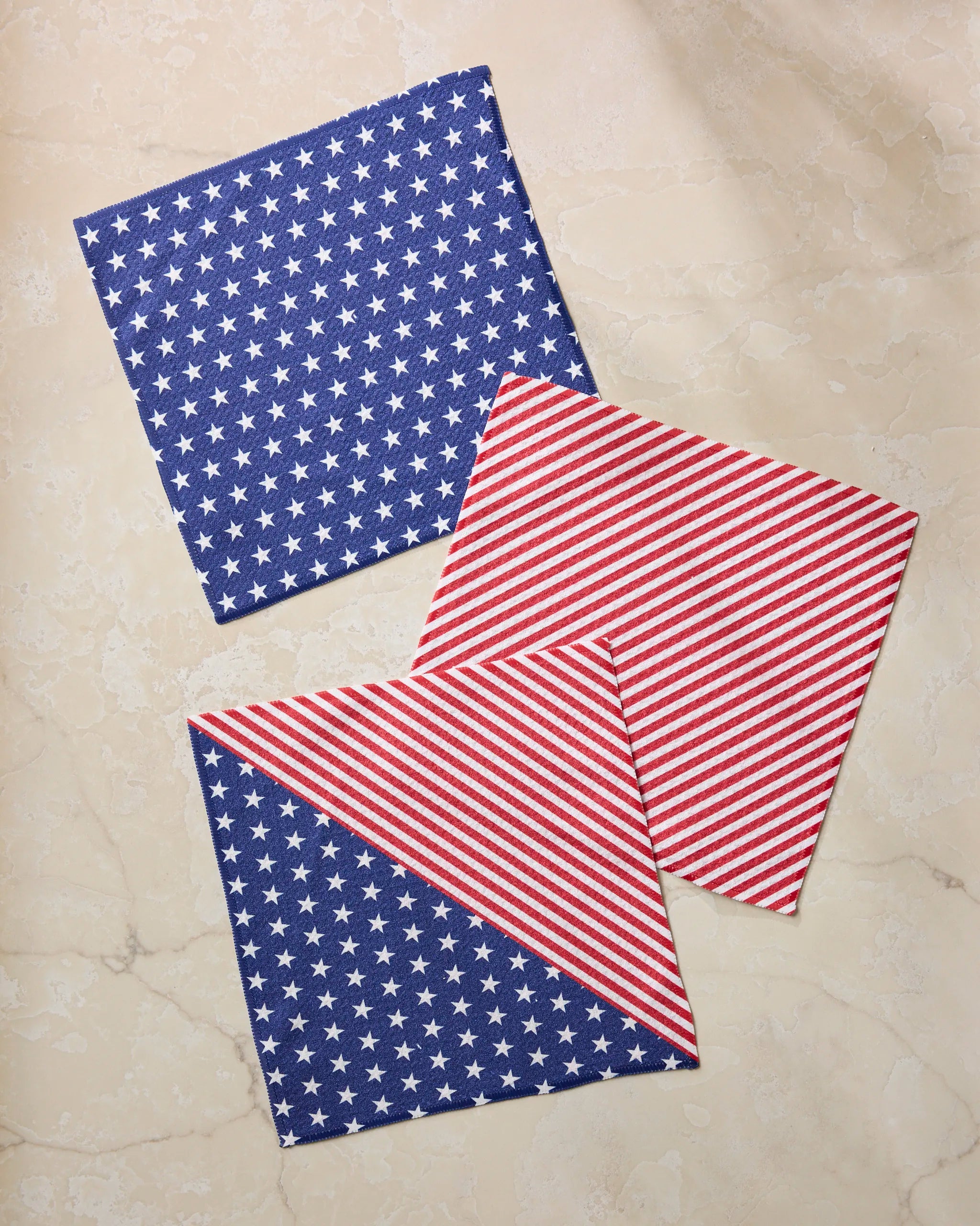 Three Liberty napkins by Geometry on a light marble surface: one with blue and white stars, one with red and white diagonal stripes, and one folded to show both designs—part of a festive recycled dishcloth set.