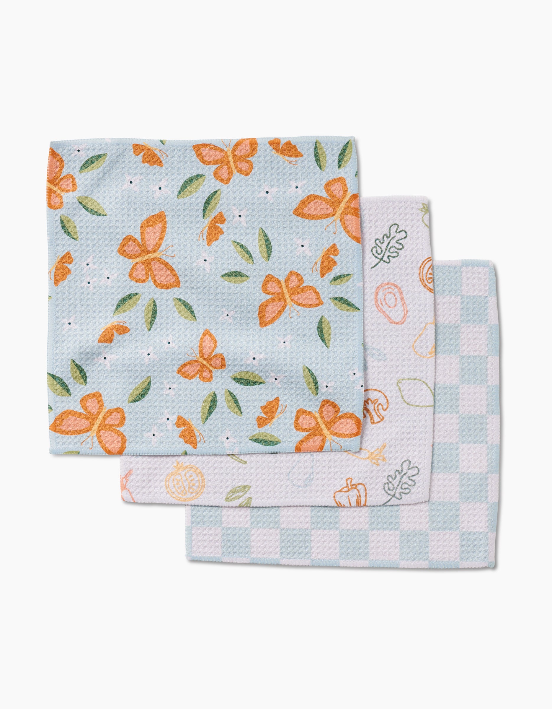 A stack of three Life is Beautiful dishcloths by Sweet Simple Vegan: the top has orange flowers and green leaves on light blue, the middle shows orange kitchen utensils on white, and the bottom is light blue and white checkered Geoweave fabric.