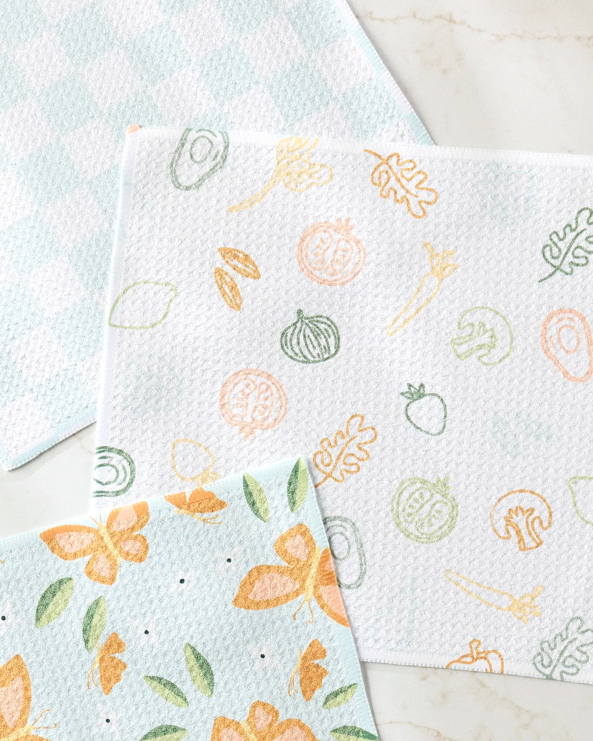 The Life is Beautiful dishcloth set by Sweet Simple Vegan includes three reusable cloths—blue checks, pastel fruits and veggies, and orange floral prints—on a light surface. Made from recycled materials for an eco-friendly clean-up.