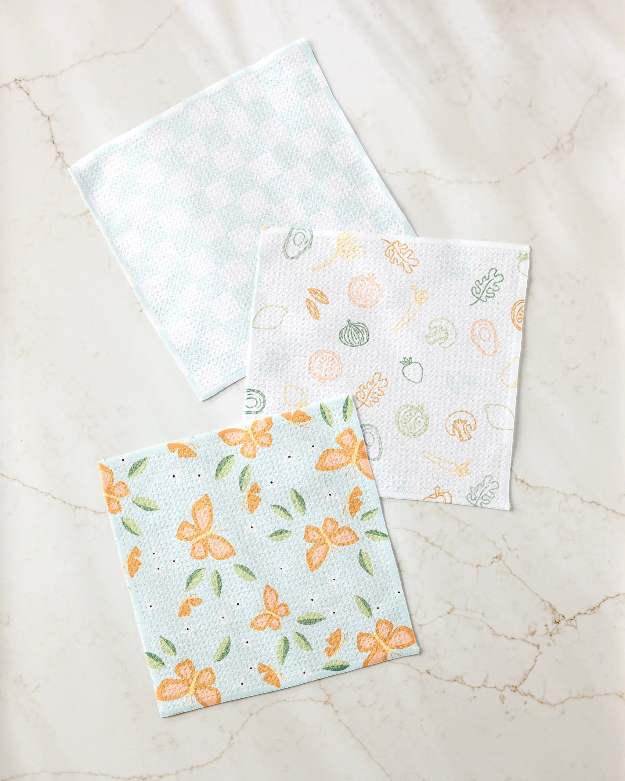 Three super-absorbent “Life is Beautiful” dishtowels by Sweet Simple Vegan are displayed on a marble surface: one with a blue gingham pattern, one with orange and green vegetable doodles, and another with an orange butterfly and floral design.