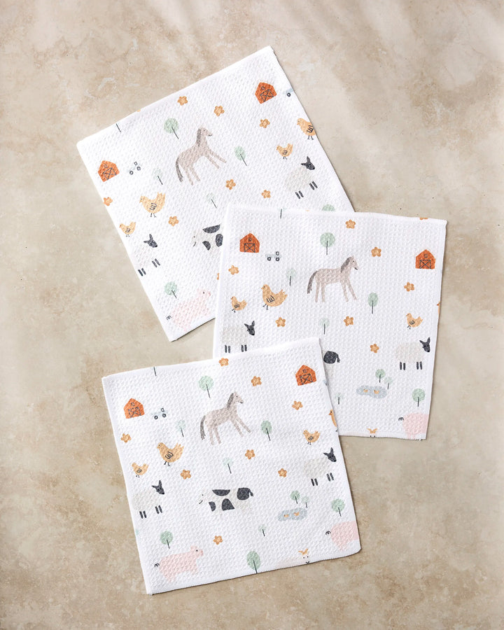 Three white paper napkins from Coit Creative's Light Farm Animals collection, featuring horses, cows, chickens, pigs, trees, flowers & barns on a beige surface—perfect paired with recycled dish towels for any farmhouse kitchen.