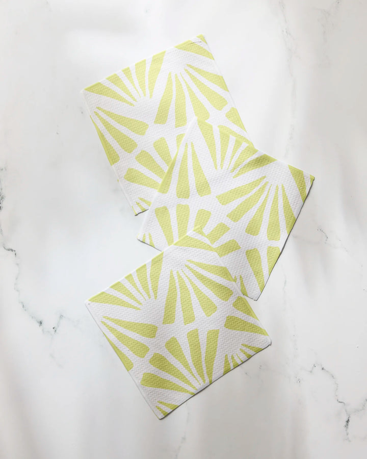 Three Geometry Lime Blossom dishtowels with yellow sunburst patterns are arranged diagonally on a white marble surface.