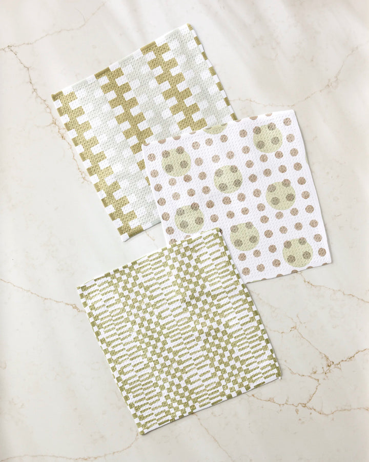 Three absorbent Lotty dishcloths by Studio Jeannot, in green and beige, lie on marble. The set features stylish geometric designs: checks, dots with circles, and zigzags.