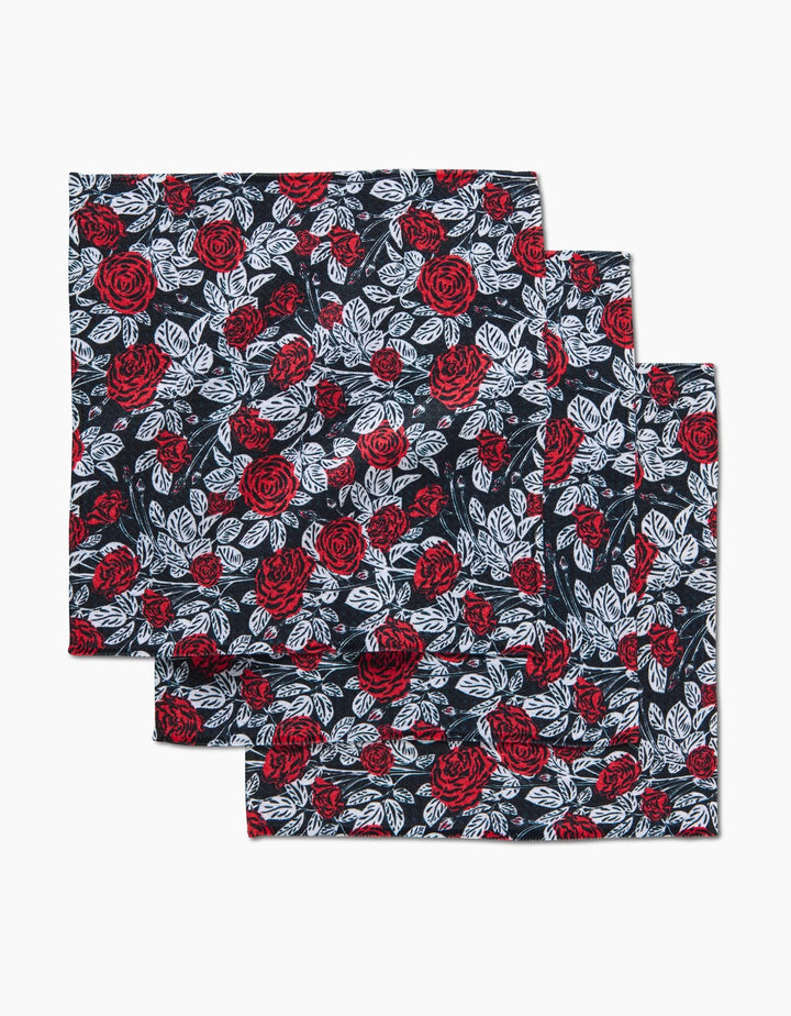 Three dark-colored Love and Roses cloth napkins by Charlie Rowan Designs, made from recycled materials, feature a repeating red rose and white leaf pattern and are neatly stacked with slight overlap on a white background.