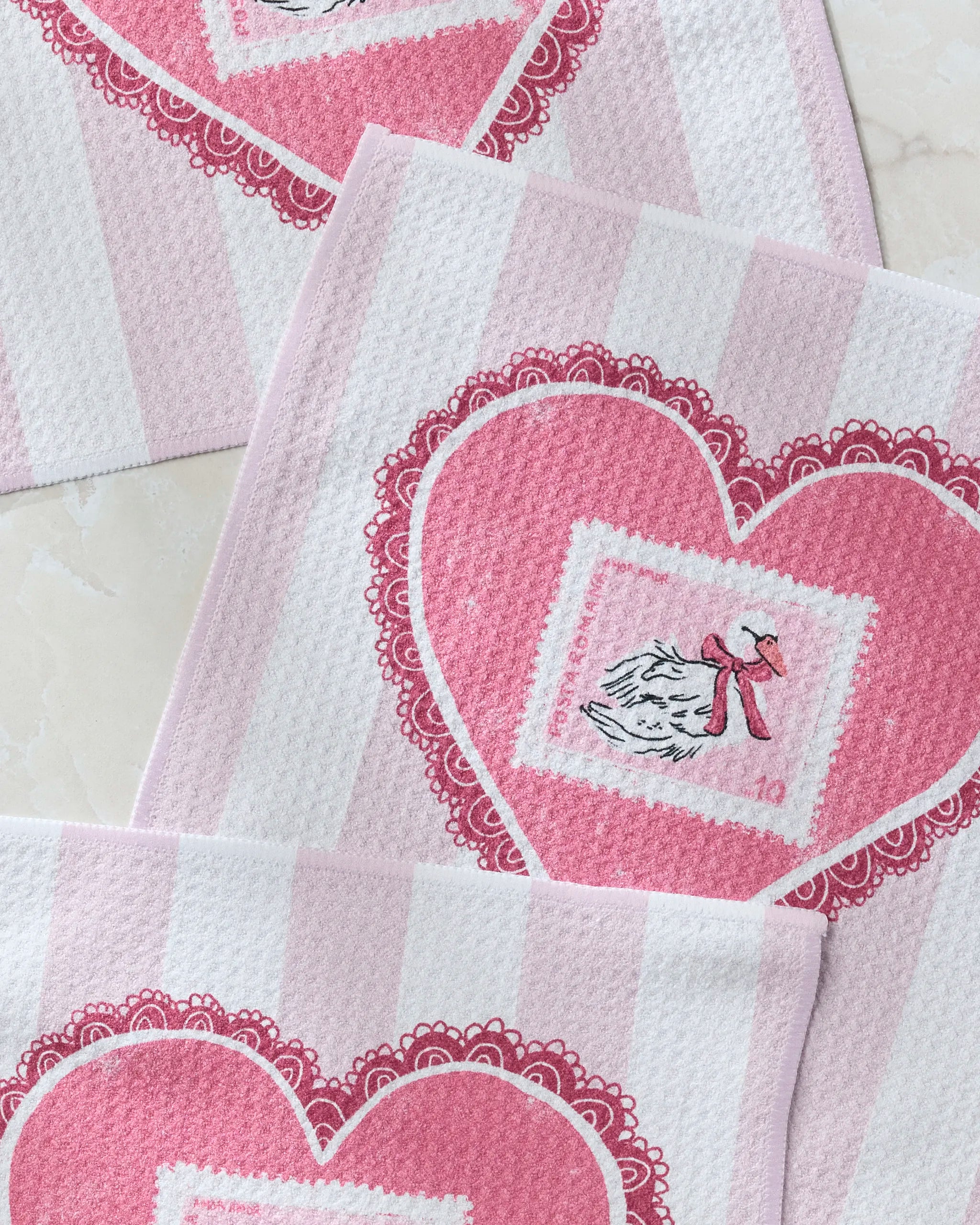 The Geometry Love and Swans dishcloth set features pink and white stripes, large pink hearts, swans in winter hats and scarves, and a lace-inspired border. Made from recycled materials, these eco-friendly dishtowels are fast-drying and charming.